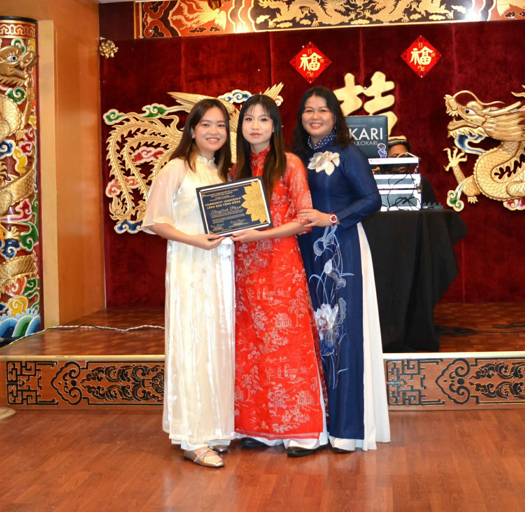 Three women in áo dài on a stage, two of them holding a framed award plaque.